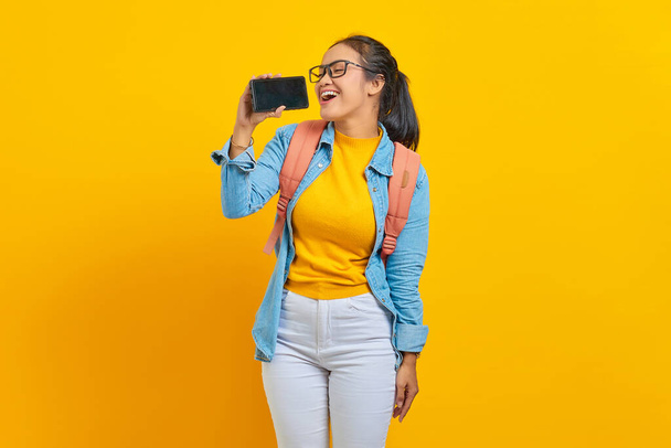 Porträt einer lächelnden jungen asiatischen Studentin in lässiger Kleidung mit Rucksack, die es genießt, mit Smartphone auf gelbem Hintergrund zu singen. Ausbildung im Hochschulkonzept - Foto, Bild