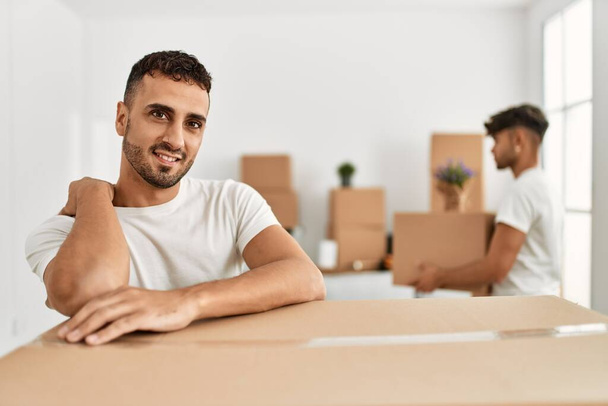 Два испаноязычных мужчины пара улыбаясь уверенно опираясь на картонную коробку в новом доме - Фото, изображение