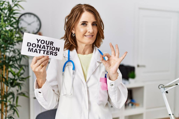 Middle age doctor woman holding your donation matters at the clinic doing ok sign with fingers, smiling friendly gesturing excellent symbol  - Foto, immagini