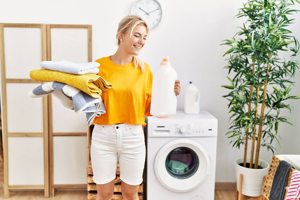 Menina loira fazendo lavanderia segurando garrafa detergente em casa. - Foto, Imagem