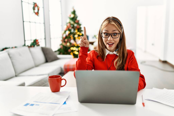 Fiatal kaukázusi lány ül az asztalon dolgozik laptoppal karácsonyfa mutatva ujjal felfelé a sikeres ötlet. kilépett és boldog volt. Egyes számú..  - Fotó, kép