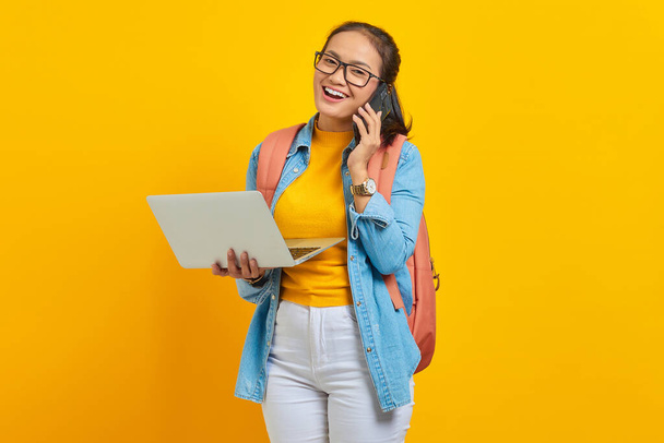 携帯電話で話すバックパックと黄色の背景に隔離されたノートパソコンを保持してカジュアルな服で陽気な若いアジアの女性の学生の肖像画。大学の概念における教育 - 写真・画像