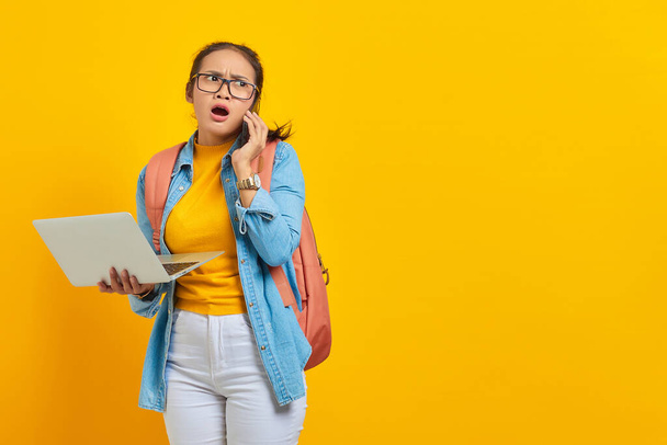 Portrait of surprised young Asian woman student in casual clothes with backpack talking on mobile phone and holding laptop isolated on yellow background. Education in college university concept - Fotoğraf, Görsel