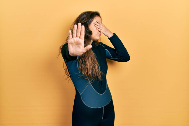 Young hispanic girl wearing diver neoprene uniform covering eyes with hands and doing stop gesture with sad and fear expression. embarrassed and negative concept.  - Фото, изображение