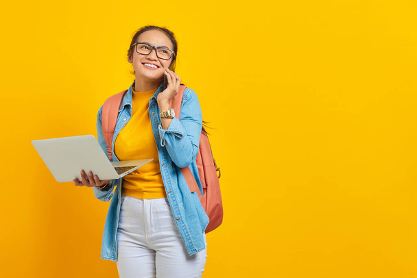 Retrato de una joven estudiante asiática alegre vestida con ropa casual con mochila hablando por teléfono móvil y sosteniendo el portátil aislado sobre fondo amarillo. Educación en concepto universitario universitario - Foto, imagen