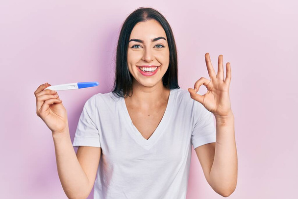 Piękna kobieta z niebieskimi oczami trzyma wynik testu ciążowego robi ok znak palcami, uśmiechając przyjazny gest doskonały symbol  - Zdjęcie, obraz