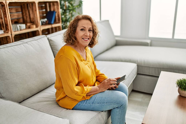 Keski-ikäinen valkoihoinen nainen käyttää älypuhelinta istuu sohvalla kotona. - Valokuva, kuva