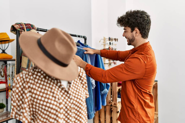 Joven cliente hispano sonriendo feliz eligiendo ropa en la tienda de ropa. - Foto, imagen
