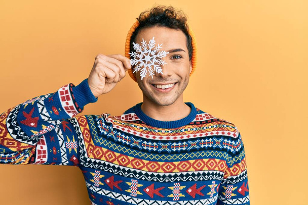 Νεαρός όμορφος άντρας που κρατάει νιφάδα χιονιού φορώντας χειμωνιάτικο πουλόβερ να δείχνει θετικός και χαρούμενος να στέκεται και να χαμογελάει με αυτοπεποίθηση να δείχνει δόντια  - Φωτογραφία, εικόνα
