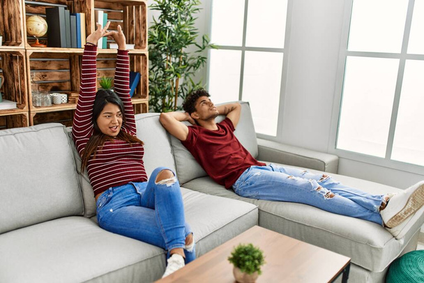 Young latin couple tired yawning and stretching arms sitting on the sofa at home. - Φωτογραφία, εικόνα