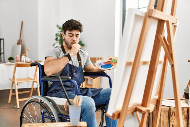 Joven hombre hispano sentado en silla de ruedas pintando en el estudio de arte sintiéndose mal y tosiendo como síntoma de frío o bronquitis. concepto de atención sanitaria.  - Foto, Imagen