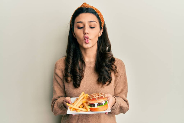 Mujer hispana joven comiendo una sabrosa hamburguesa clásica haciendo cara de pez con boca y ojos entrecerrados, loca y cómica.  - Foto, imagen