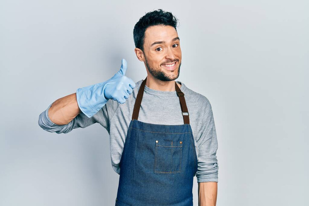 Young hispanic man wearing apron and gloves smiling happy and positive, thumb up doing excellent and approval sign  - Photo, Image