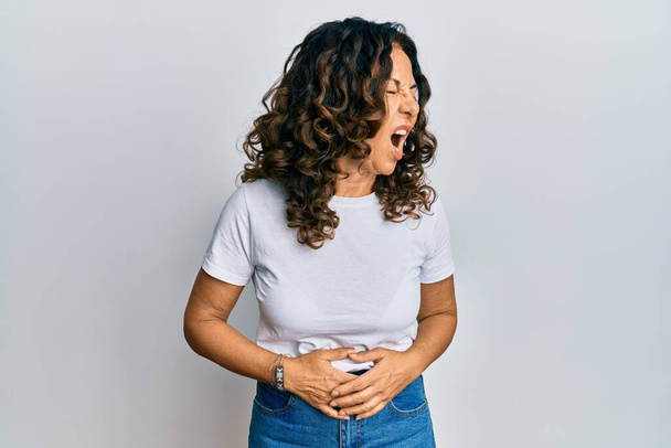 中年のヒスパニック系の女性が胃の上に手でカジュアルな白いTシャツを着て吐き気、痛みを伴う病気の気分が悪い。キャッシュ・コンセプト.  - 写真・画像