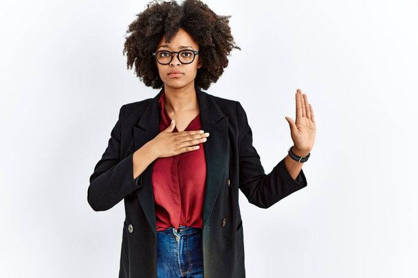 Donna afro-americana con capelli afro che indossa giacca da lavoro e occhiali che giurano con mano sul petto e palmo aperto, facendo un giuramento di fedeltà  - Foto, immagini