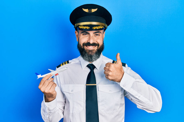 Jeune homme hispanique portant uniforme de pilote tenant jouet avion souriant heureux et positif, pouce levé faire excellent et signe d'approbation  - Photo, image