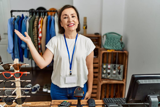 Beautiful middle age hispanic woman working as manager at retail boutique smiling cheerful presenting and pointing with palm of hand looking at the camera.  - Photo, Image