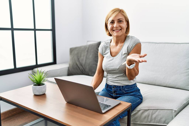 Femme blonde du moyen âge utilisant un ordinateur portable à la maison pointant de côté avec les mains ouvertes paumes montrant l'espace de copie, présentant la publicité souriant heureux excité  - Photo, image