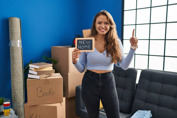 Jeune femme hispanique tenant un tableau noir avec un nouveau texte à la maison souriant avec une idée ou une question pointant du doigt avec un visage heureux, numéro un  - Photo, image