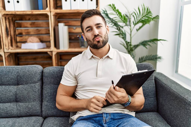 Handsome hispanic man holding clipboard working at psychology clinic looking at the camera blowing a kiss on air being lovely and sexy. love expression.  - Φωτογραφία, εικόνα