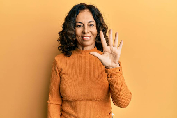 Mujer hispana de mediana edad vistiendo ropa casual mostrando y señalando con los dedos el número cinco mientras sonríe confiada y feliz.  - Foto, imagen