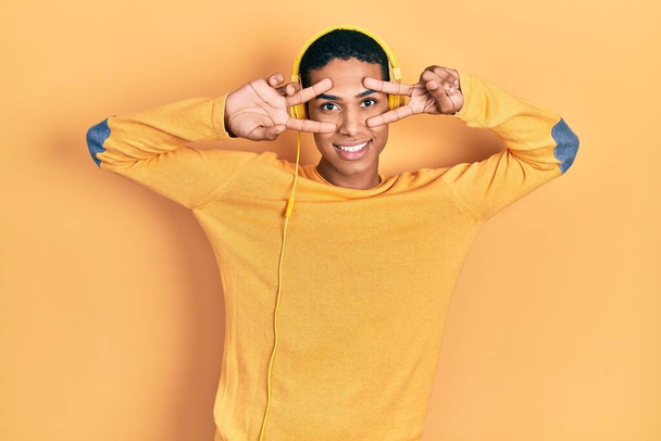 Young african american guy listening to music using headphones doing peace symbol with fingers over face, smiling cheerful showing victory  - Foto, immagini