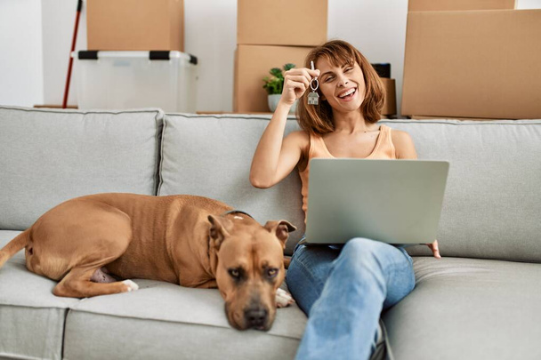 Jovem mulher caucasiana tendo chamada de vídeo e segurando a chave sentada no sofá com o cão em casa - Foto, Imagem
