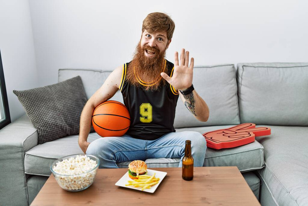 Καυκάσιος άντρας με μακριά γενειάδα κρατώντας μπάλα του μπάσκετ ζητωκραυγάζει τηλεοπτικό παιχνίδι παραίτηση λέγοντας γεια χαρούμενος και χαμογελαστός, φιλική χειρονομία καλωσορίσματος  - Φωτογραφία, εικόνα
