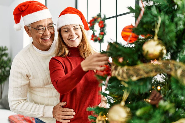 Pareja hispana de mediana edad sonriendo feliz decorando el árbol de Navidad en casa. - Foto, imagen
