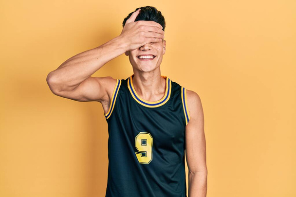 バスケットボールの制服を着た若いヒスパニック系の男性が笑顔で、驚きのために目を覆う顔を手で笑っています。視覚障害の概念.  - 写真・画像
