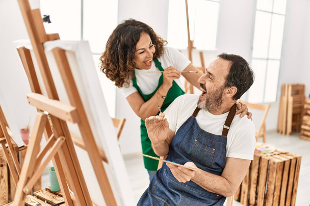 Δύο μαθητές μέσης ηλικίας χαμογελώντας ευτυχισμένοι παίζοντας με ζωγραφική πινέλο ο ένας στον άλλο στη Σχολή Καλών Τεχνών. - Φωτογραφία, εικόνα