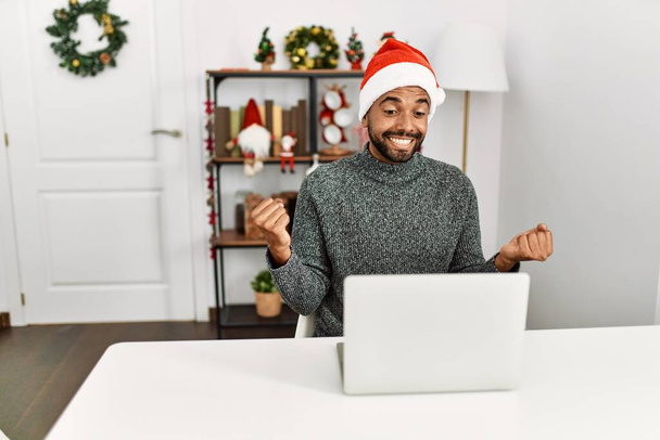 Young hispanic man with beard wearing christmas hat using laptop screaming proud, celebrating victory and success very excited with raised arms  - Photo, Image