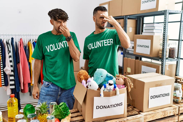 Young gay couple wearing volunteer t shirt at donations stand smelling something stinky and disgusting, intolerable smell, holding breath with fingers on nose. bad smell  - Photo, Image