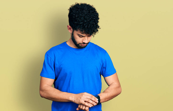 Młody arabski mężczyzna z brodą w luźnej niebieskiej koszulce sprawdzający czas na zegarku, zrelaksowany i pewny siebie  - Zdjęcie, obraz
