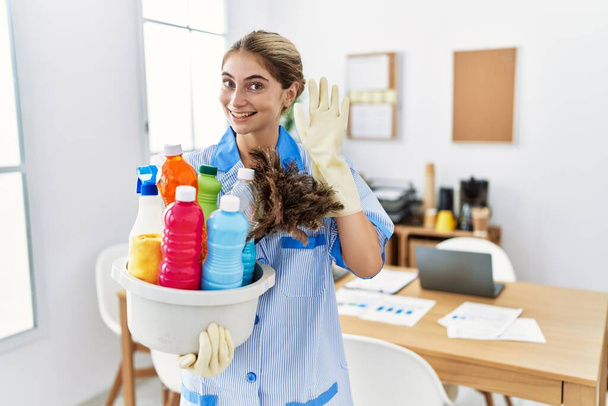 Young blonde woman wearing cleaner uniform holding cleaning products waiving saying hello happy and smiling, friendly welcome gesture  - Photo, Image