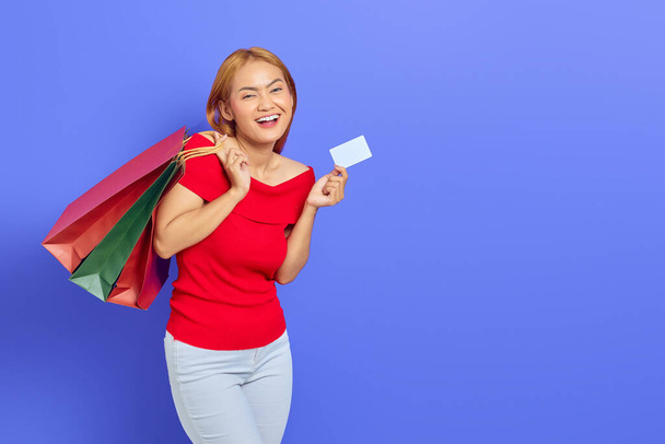 Веселая красивая азиатская женщина в красном платье, держащая сумки с покупками и кредитные карты изолированы на фиолетовом фоне - Фото, изображение