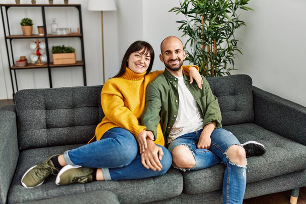 Young hispanic couple smiling happy and hugging sitting on the sofa at home. - 写真・画像