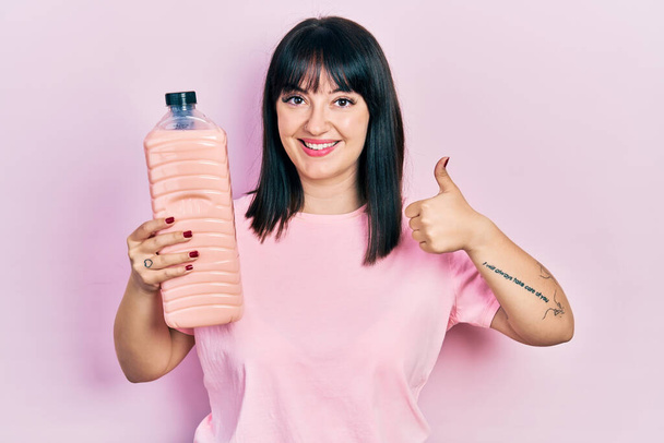 Mujer hispana joven sosteniendo botella de detergente sonriendo feliz y positivo, pulgar hacia arriba haciendo excelente y signo de aprobación  - Foto, imagen