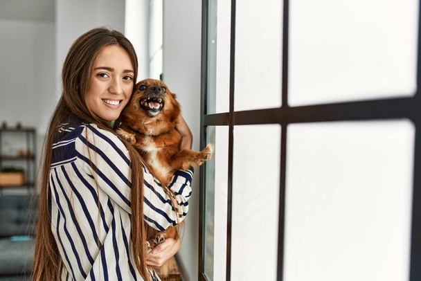 Νεαρή ισπανόφωνη γυναίκα χαμογελά με αυτοπεποίθηση αγκαλιάζει το σκυλί στέκεται στο σπίτι - Φωτογραφία, εικόνα
