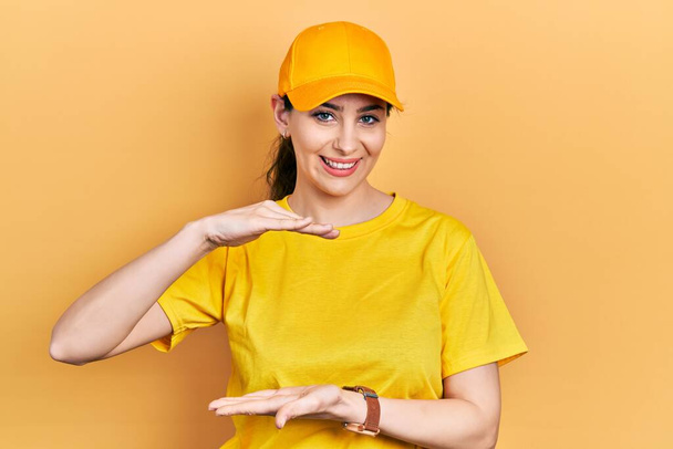 Young hispanic woman wearing delivery uniform and cap gesturing with hands showing big and large size sign, measure symbol. smiling looking at the camera. measuring concept.  - Photo, Image