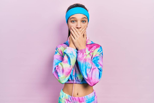 Young brunette girl wearing gym clothes and headphones shocked covering mouth with hands for mistake. secret concept.  - Photo, Image