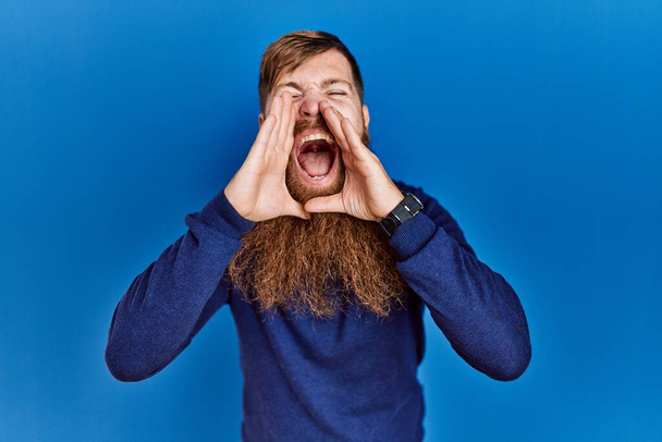 Homme rousse à longue barbe portant un pull bleu décontracté sur fond bleu criant à haute voix avec les mains sur la bouche  - Photo, image