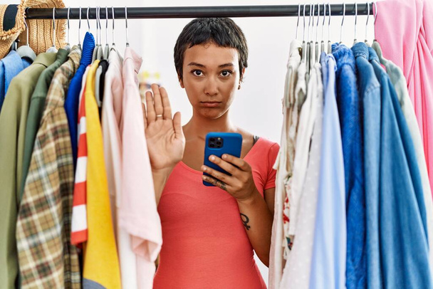 Jeune femme hispanique aux cheveux courts cherchant des vêtements sur un porte-vêtements à l'aide d'un smartphone à la main ouverte faisant signe d'arrêt avec une expression sérieuse et confiante, geste de défense  - Photo, image