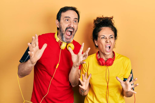 Middle age couple of hispanic woman and man wearing sportswear and arm band crazy and mad shouting and yelling with aggressive expression and arms raised. frustration concept.  - 写真・画像