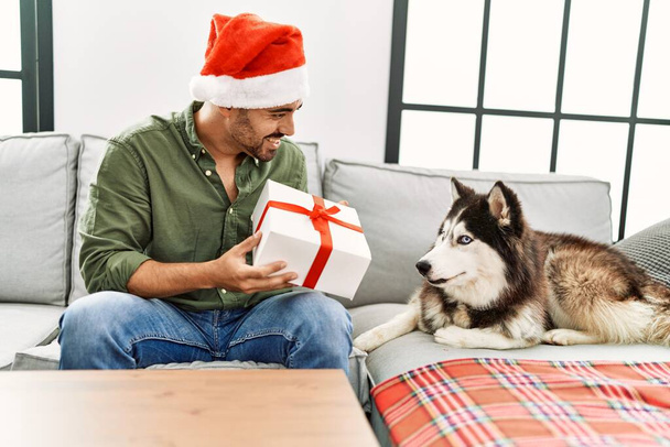 Young hispanic man holding gift sitting on sofa with dog by christmas decor at home - Photo, Image