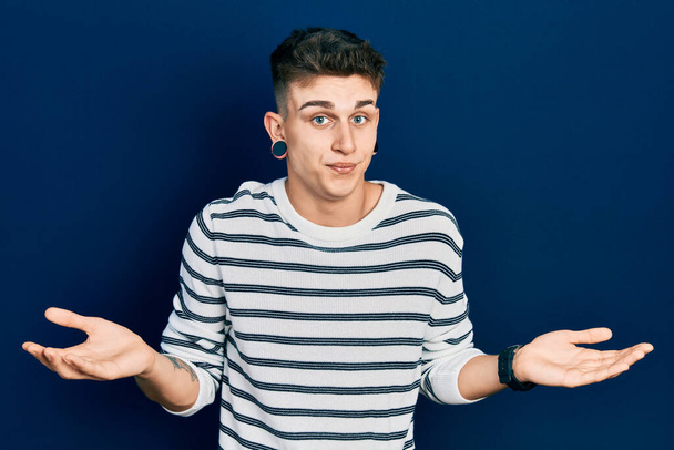 Young caucasian boy with ears dilation wearing casual striped shirt clueless and confused expression with arms and hands raised. doubt concept.  - Photo, Image