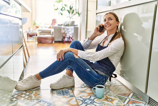 Jovem loira falando no smartphone sentado no chão na cozinha - Foto, Imagem