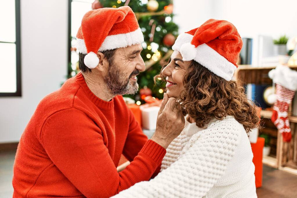 Pareja hispana de mediana edad con sombrero de navidad sentado en el suelo y abrazándose en casa. - Foto, Imagen