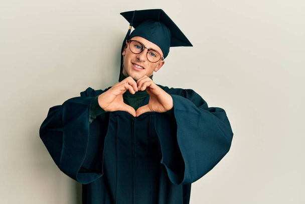 Young caucasian man wearing graduation cap and ceremony robe smiling in love doing heart symbol shape with hands. romantic concept.  - Fotó, kép