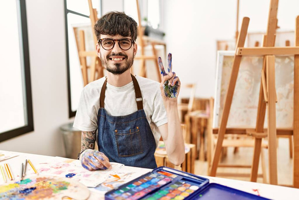 Spanyol férfi szakállal a művészeti stúdióban, a kettes számú ujjal mutogatva, miközben magabiztosan és boldogan mosolyog..  - Fotó, kép